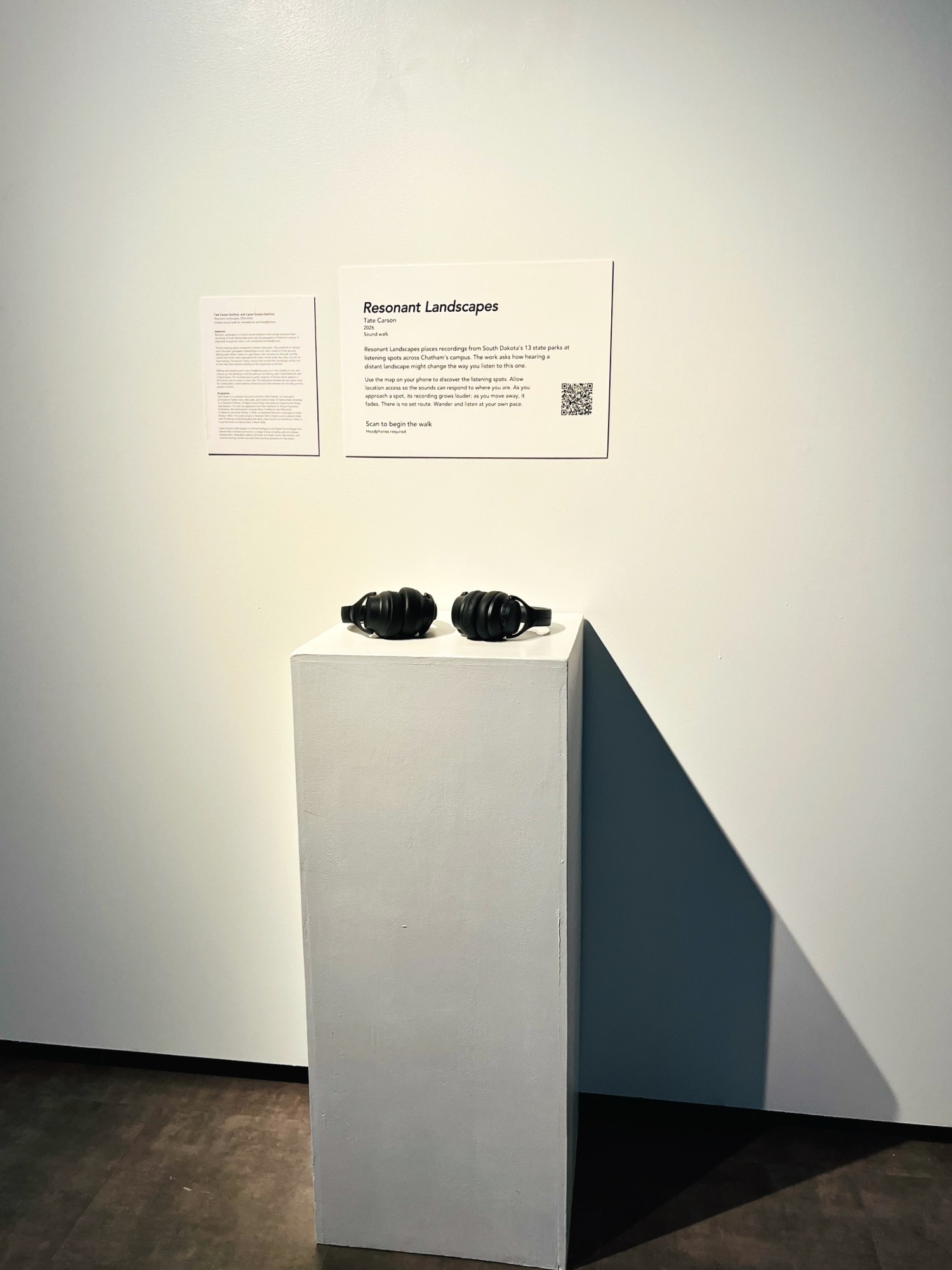 Sound+ Installations, Susan Bergman Gurrentz '56 Art Gallery, Chatham University, 2026 The Resonant Landscapes wall text, with its QR code, hung on a white gallery wall beside a smaller printed statement, above a white plinth holding a pair of black headphones.